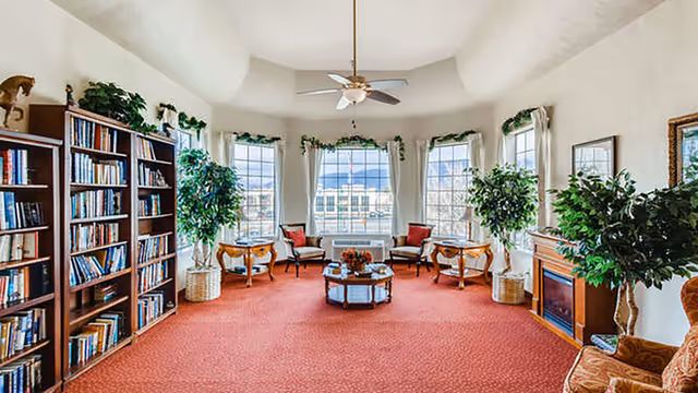 A bright and spacious living room with large windows letting in natural light. The room features a red carpet, a ceiling fan, several potted plants, a wooden bookshelf filled with books, a fireplace, and multiple chairs arranged around a central coffee table with a flower arrangement.