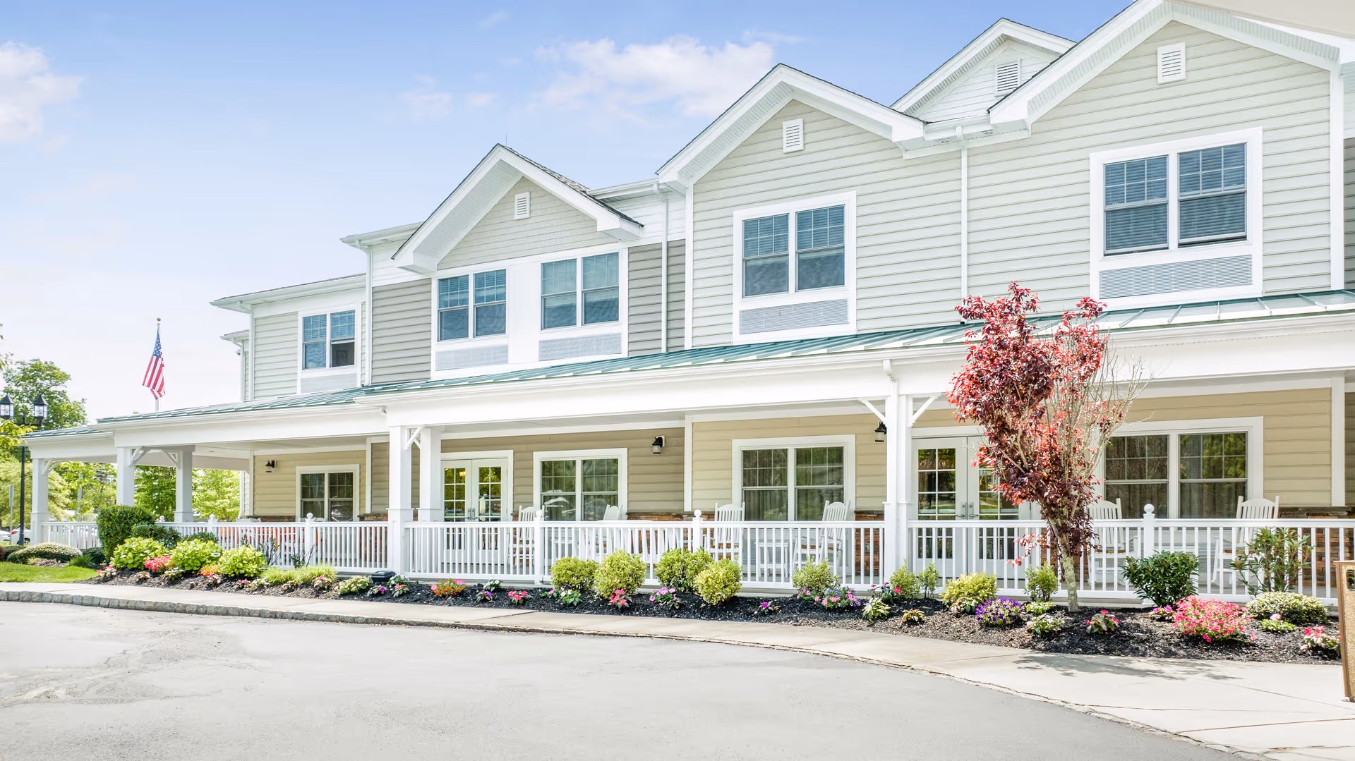 Exterior view of a two-story senior living facility building with beige siding, white trim, and a covered porch with white railings and rocking chairs. There are landscaped flower beds with shrubs and colorful flowers in front of the porch, and an American flag is visible on the left side. The sky is clear and blue.