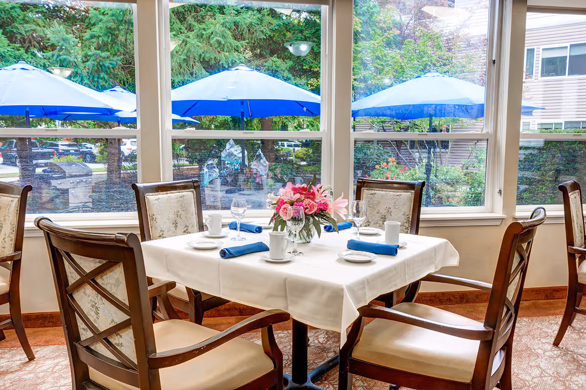 A dining table set for four with white tablecloth, blue napkins, white plates, cups, and wine glasses. A vase with pink and red flowers is in the center. The table is surrounded by wooden chairs with cushioned seats and floral upholstery. Large windows behind the table show blue patio umbrellas and greenery outside.