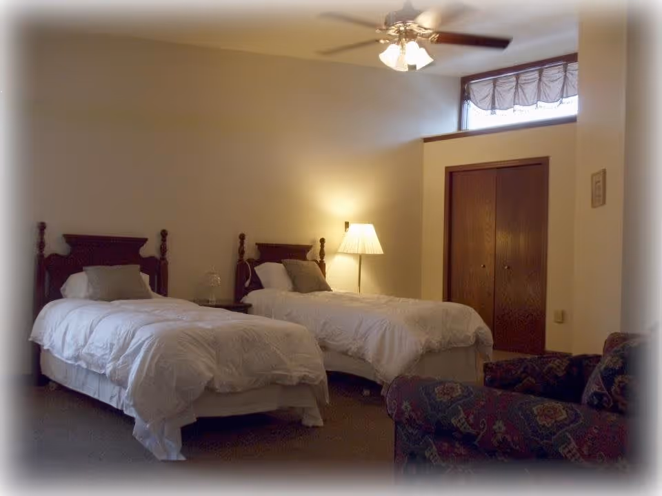 A cozy bedroom with two twin beds featuring wooden headboards, white bedding, and gray pillows. A floor lamp is positioned between the beds, casting warm light. There is a ceiling fan with lights above, a wooden closet with double doors, and a patterned armchair in the foreground.