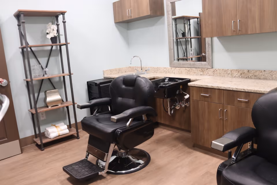 Interior of a salon room with two black salon chairs, a black hair washing sink, wooden cabinets with granite countertops, a mirror on the wall, and a metal shelving unit holding towels and decorative items.