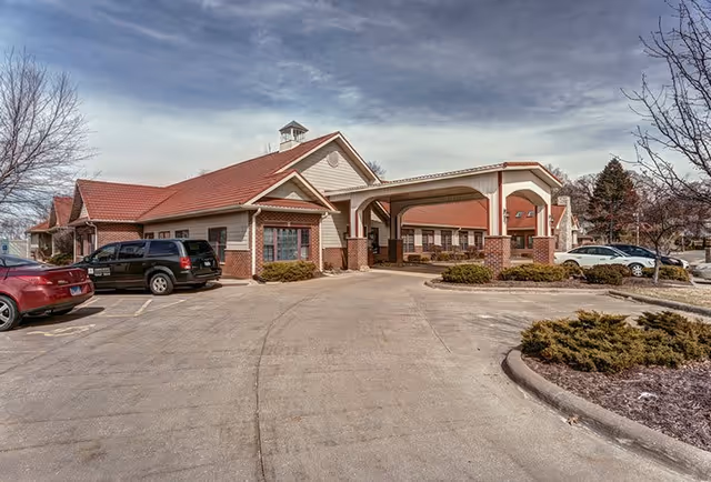 Exterior view of a senior living facility building with a covered entrance, red roof, and brick and beige siding. Several parked cars are visible in the parking lot, along with some leafless trees and shrubs under a partly cloudy sky.