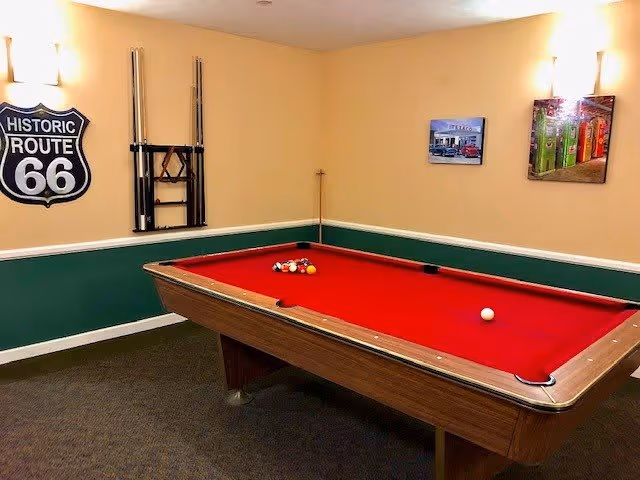 A room with a red felt pool table set up for a game, with a rack of pool cues mounted on the wall. The walls are painted beige and green with white trim, and there are three wall decorations including a Historic Route 66 sign and two framed pictures.
