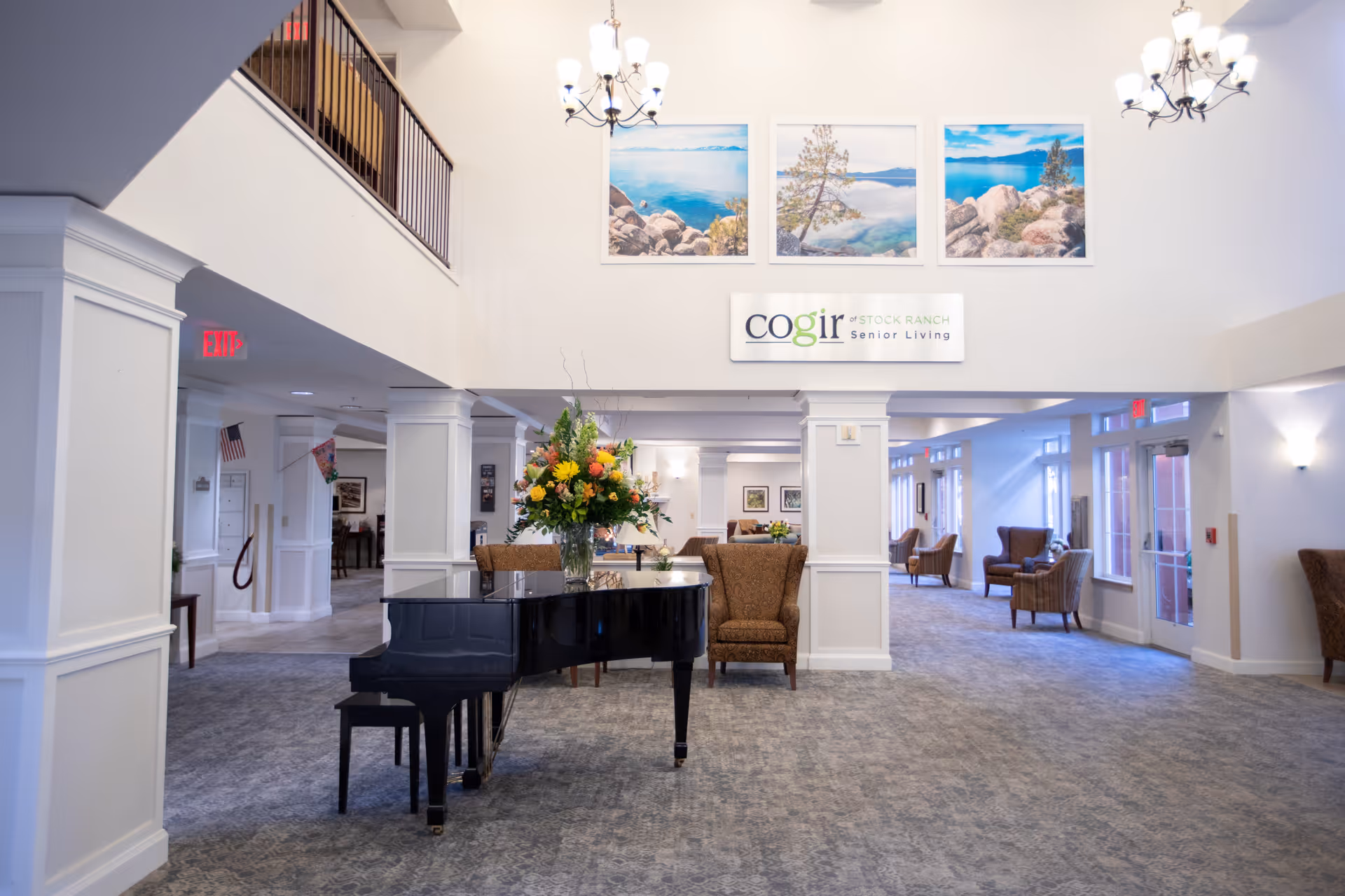 Spacious and well-lit senior living facility common area with a black grand piano adorned with a large floral arrangement, comfortable armchairs, and decorative landscape paintings on the wall. The area features high ceilings, chandeliers, and a carpeted floor with white pillars and large windows allowing natural light.