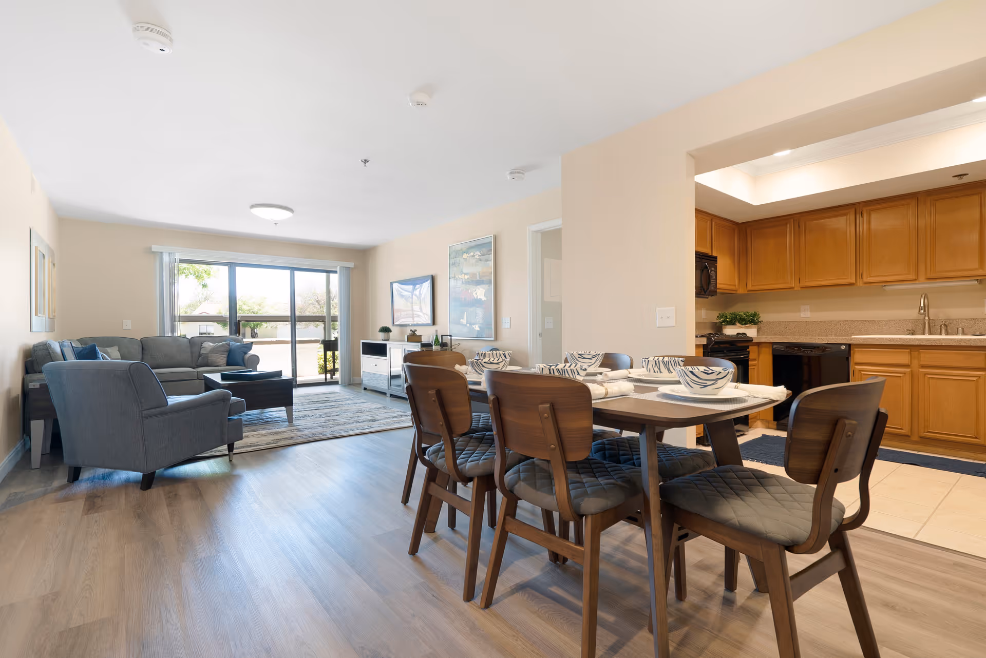 A bright and spacious living and dining area in a senior living facility. The dining area features a wooden table set with six chairs and tableware. Adjacent to the dining area is a kitchen with wooden cabinets and modern appliances. The living room has a gray sofa, an armchair, a coffee table, and a large window with sliding glass doors leading to an outdoor space.