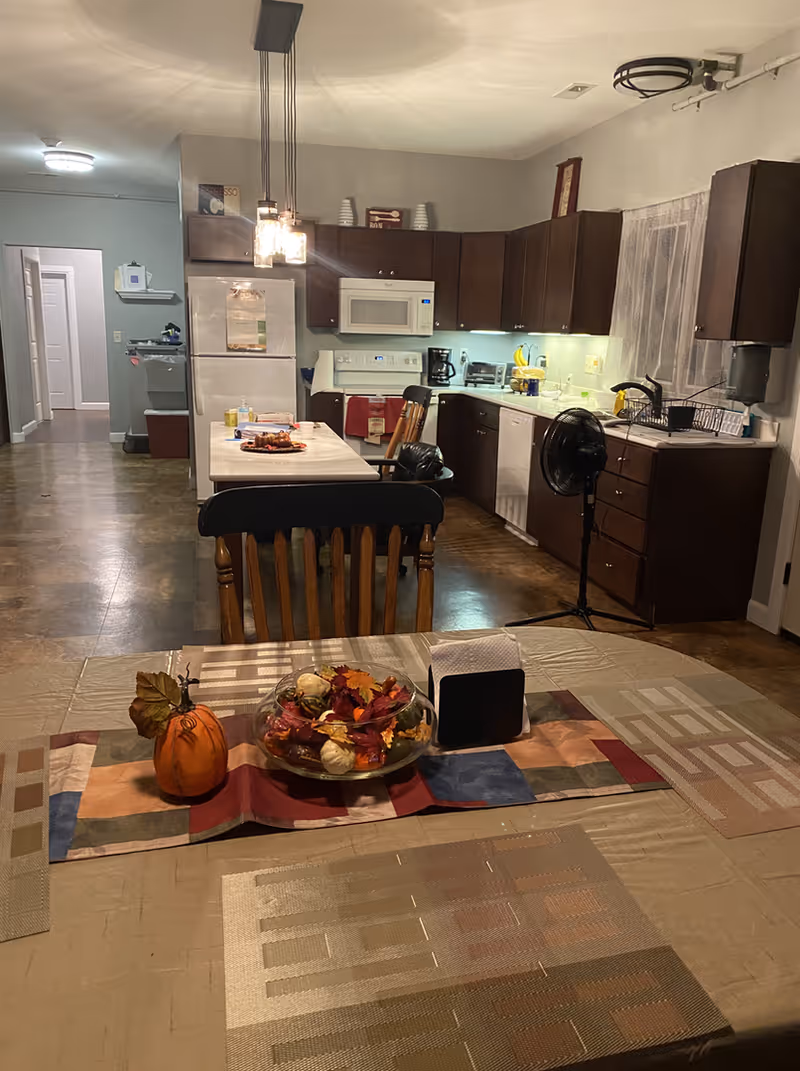 Interior view of a communal kitchen and dining area with a table centerpiece, island, refrigerator, cabinets, and chairs.