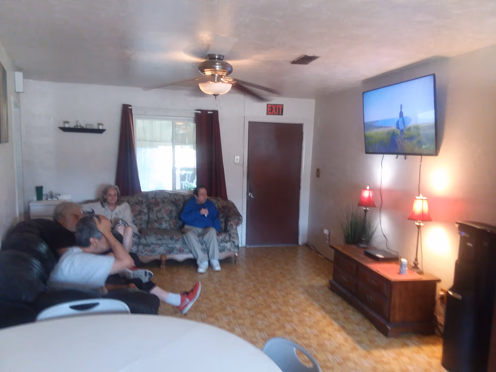 A living room with four elderly people sitting on two couches watching a wall-mounted television. The room has a ceiling fan, a door with an exit sign above it, two red lamps on a wooden cabinet, and a small black refrigerator. The floor has a patterned design, and there is a round table with chairs in the foreground.