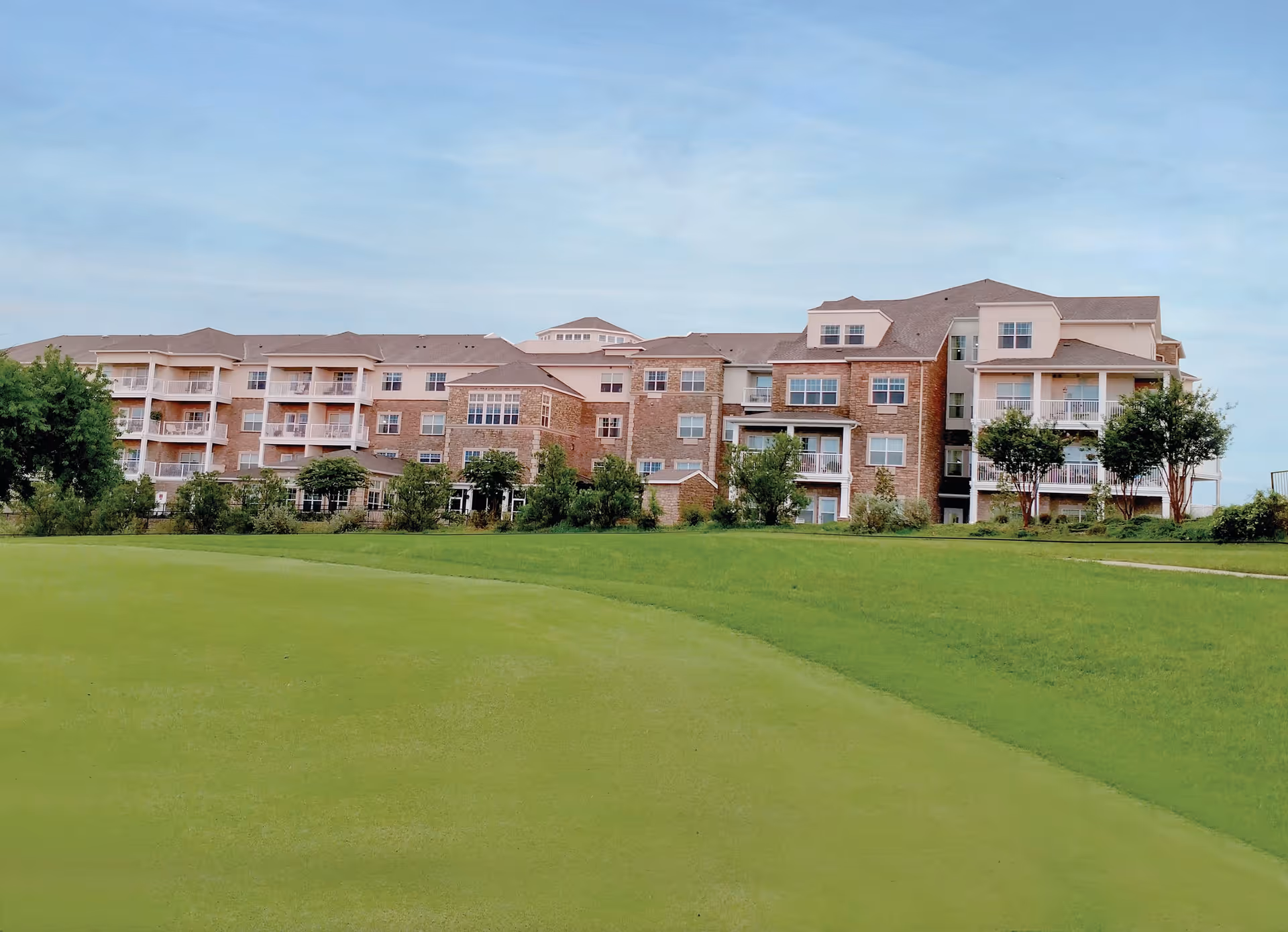 Exterior view of a large multi-story senior living facility building with balconies, surrounded by green grass and trees under a clear blue sky.