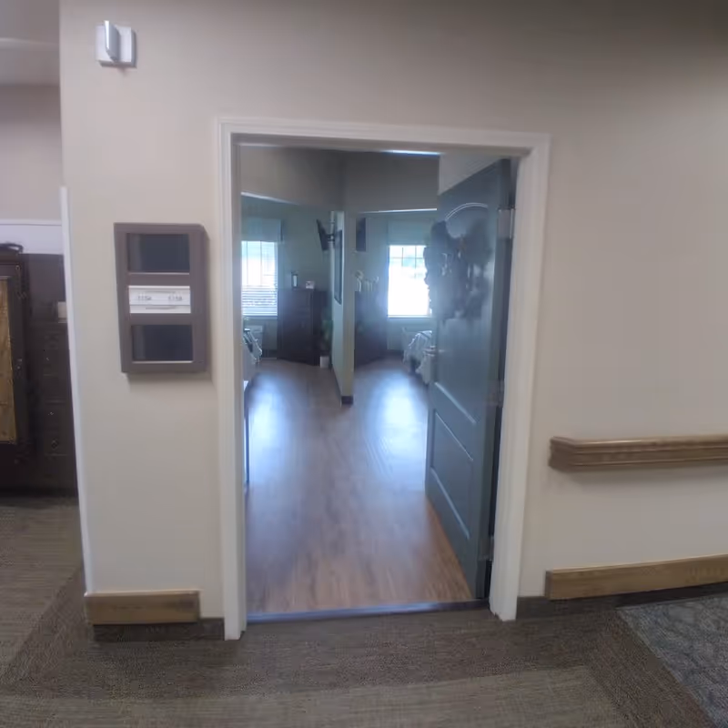 View through an open doorway into a senior living facility room with wooden flooring, a bed, a dresser, and a window with blinds letting in natural light. The hallway outside the room has carpeted flooring and beige walls with wooden trim and a handrail.