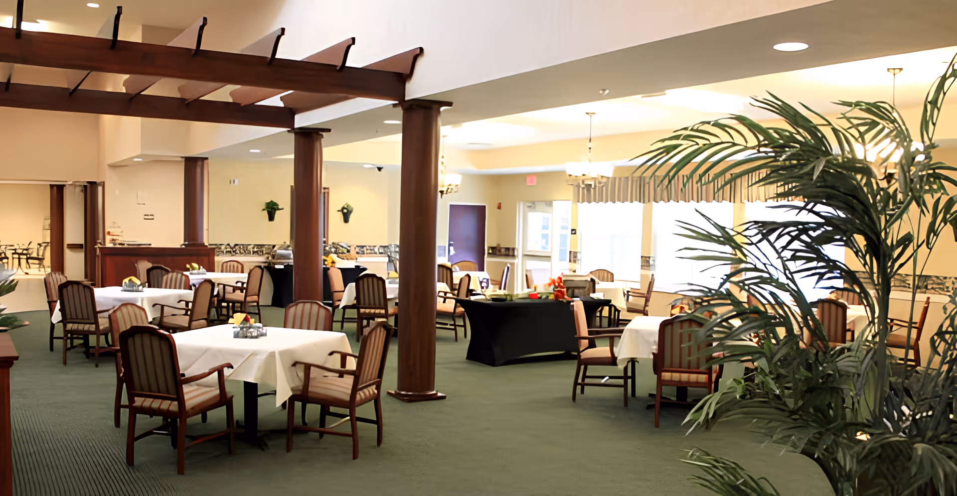 Spacious dining room with tables covered in white tablecloths, wooden columns and chairs, and large indoor plants.