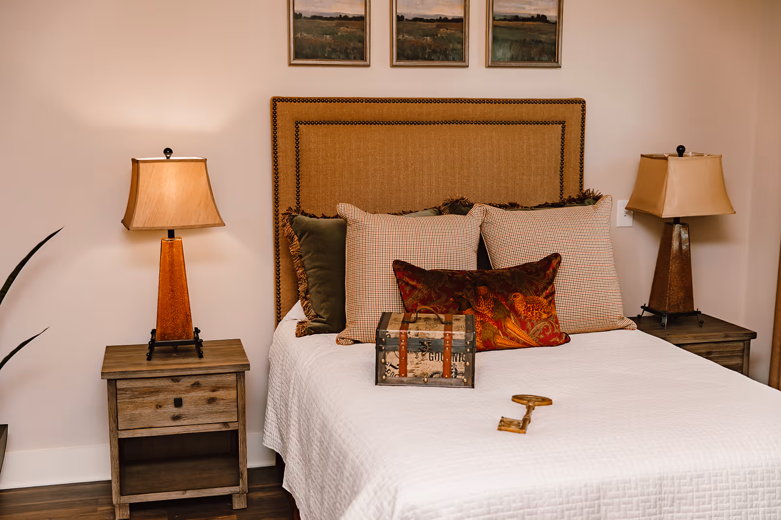 A cozy bedroom with a bed featuring a beige upholstered headboard and multiple decorative pillows. There are two wooden nightstands on either side of the bed, each with a lamp that has a beige lampshade. On the bed, there is a small decorative chest and a large vintage-style key. Above the bed, three framed landscape paintings are hung on the wall.