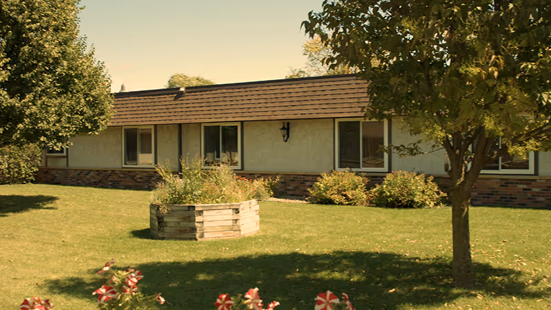 Single-story building with beige walls and a brown shingled roof, surrounded by a green lawn with trees, bushes, and a raised wooden planter box filled with plants. Red and white flowers are visible in the foreground.
