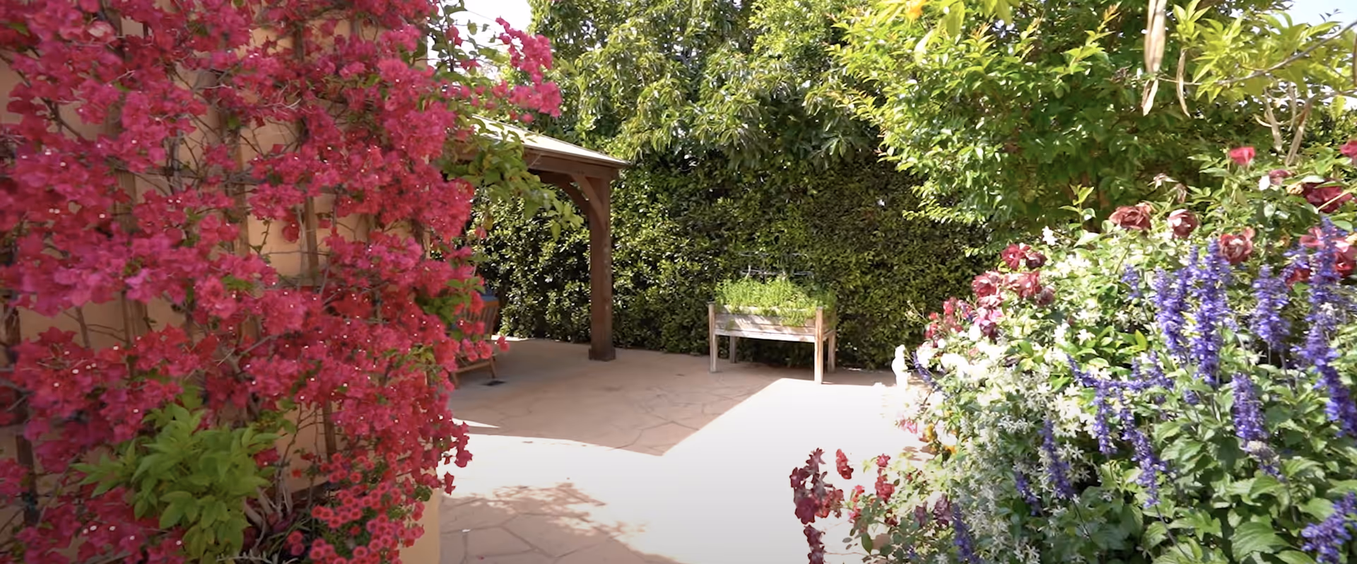 A sunny outdoor patio area surrounded by lush greenery and vibrant flowers including pink, purple, and white blooms. There is a wooden pergola with a chair underneath and a raised garden bed with herbs or plants. The space is paved with stone tiles and enclosed by tall hedges.