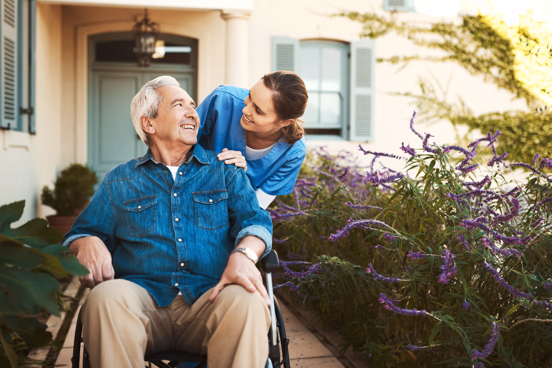 A smiling elderly man in a wheelchair is outdoors next to purple flowering plants, being gently supported by a female caregiver in blue scrubs who is leaning towards him with a caring expression.