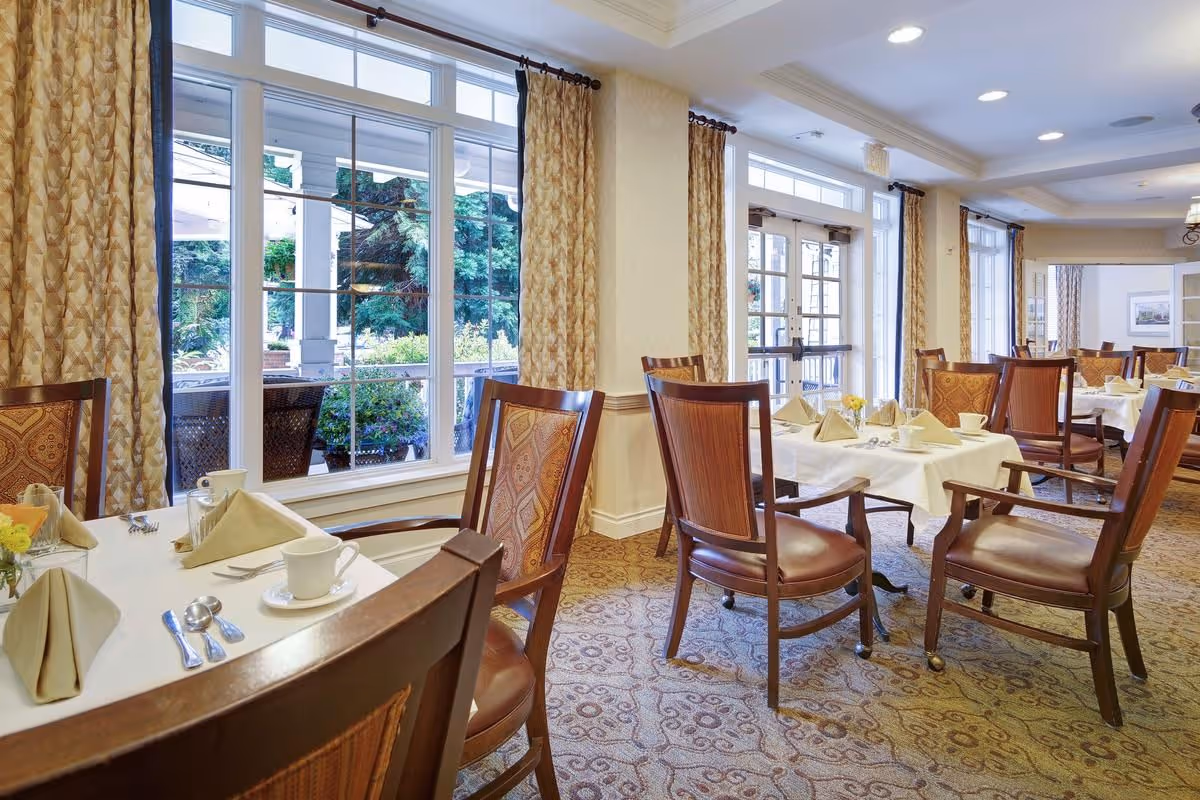 A bright dining room with several tables covered in white tablecloths, set with beige napkins, cups, and silverware. The room has large windows with patterned curtains, allowing natural light to fill the space. Wooden chairs with cushioned seats and backs surround the tables. Outside the windows, greenery and outdoor seating are visible.