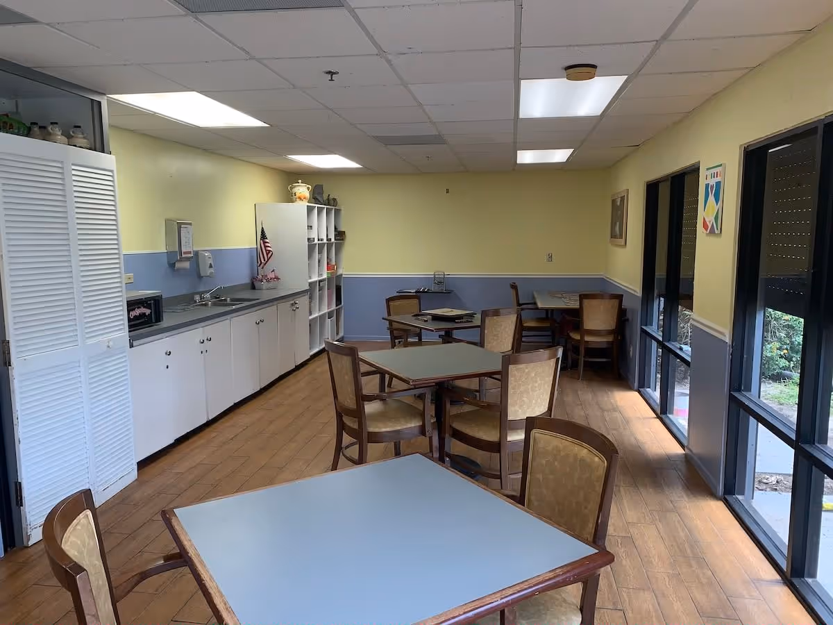 Small dining/activity room with multiple square tables and chairs, a kitchenette and storage along the left wall, and large windows on the right.