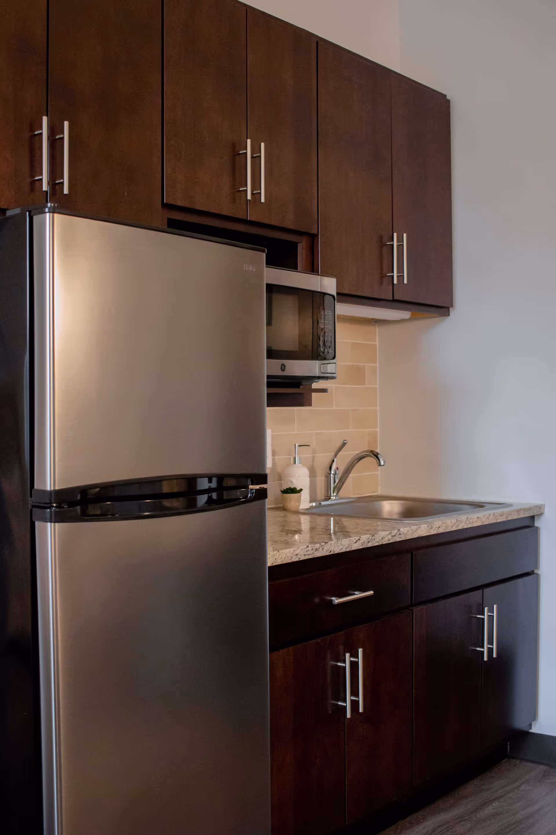 A kitchen area with dark brown cabinets, a stainless steel refrigerator, a microwave mounted above the countertop, a sink with a faucet, and a soap dispenser next to a small potted plant. The backsplash is made of beige tiles.