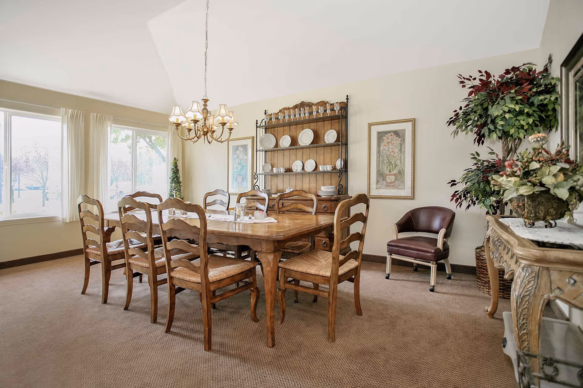 A bright dining room with a large wooden table surrounded by eight chairs, a chandelier, china hutch, framed art, and decorative plants.