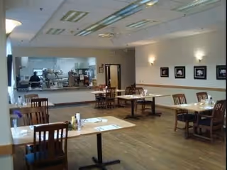 A dining room with several wooden tables and chairs arranged neatly. The room has wooden flooring, framed pictures on the wall, and a large window-like opening showing a kitchen area in the background. The lighting is soft with wall-mounted lamps and ceiling lights.