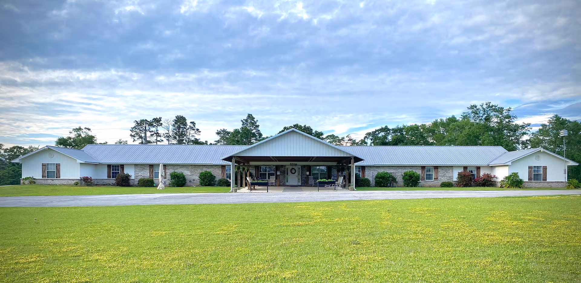Wide exterior view of a single-story senior living facility building with a metal roof, a covered entrance, and surrounded by green grass and trees under a partly cloudy sky.