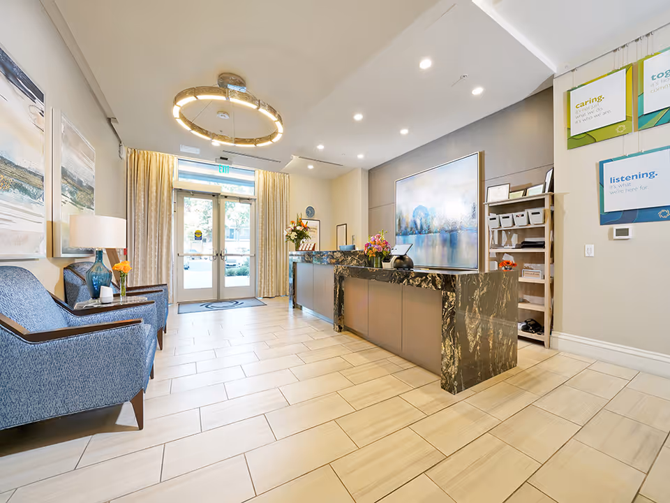 Bright reception lobby with a marble front desk, blue seating, artwork, and double glass entry doors.
