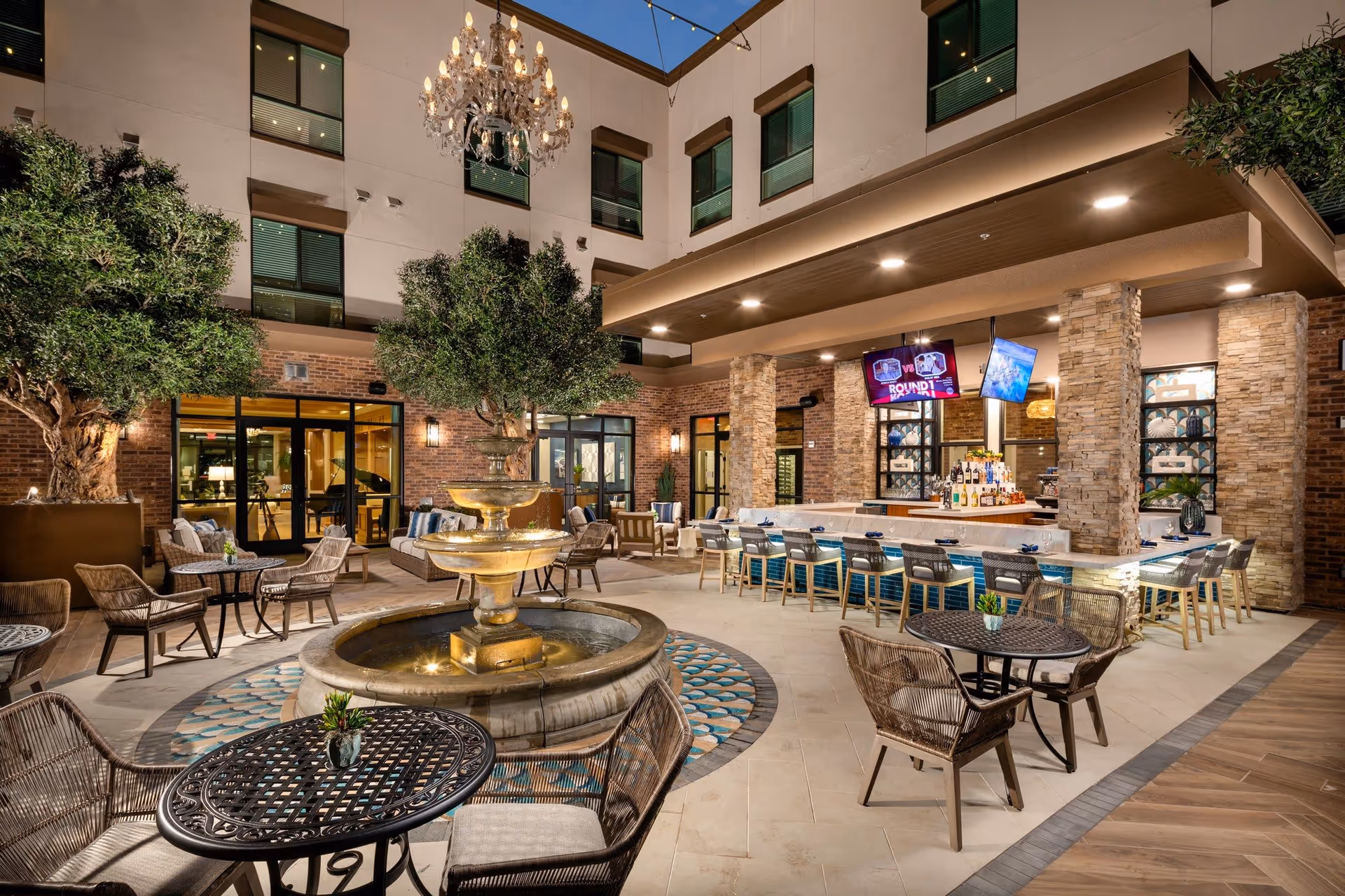 Outdoor courtyard area at Clearwater Newport Beach featuring a central water fountain surrounded by round tables and wicker chairs. There is a bar with high stools under a covered patio supported by stone columns. Two televisions are mounted above the bar. The courtyard is decorated with trees, string lights, and a chandelier hanging above the fountain.