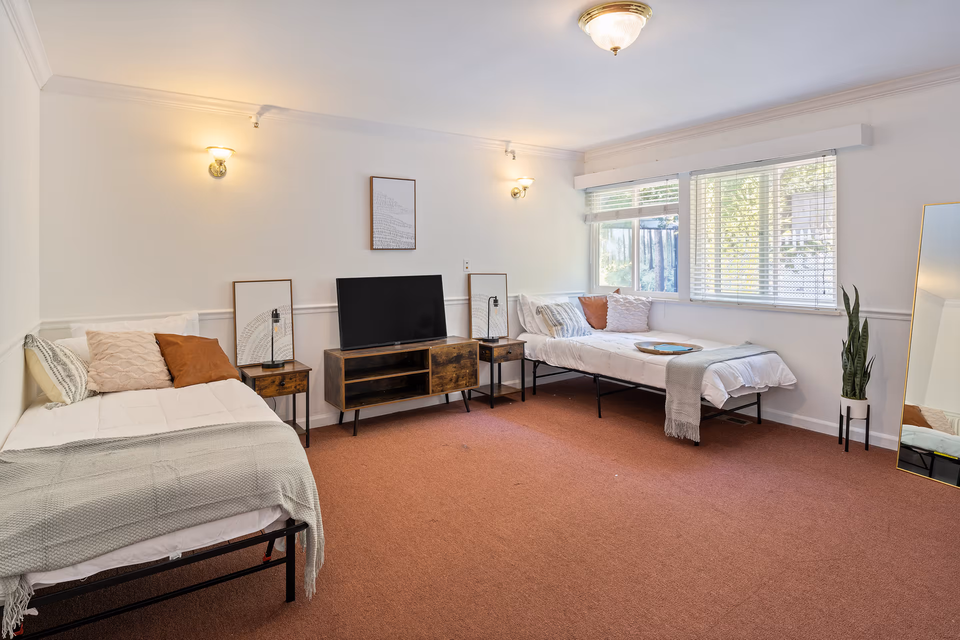 A bright and spacious bedroom with two single beds, each adorned with pillows and blankets. Between the beds is a wooden TV stand with a flat-screen TV and two small side tables with lamps and framed artwork. A large window with blinds lets in natural light, and a tall mirror and a potted plant are placed near the window. The room has white walls, carpeted floor, and ceiling lights.