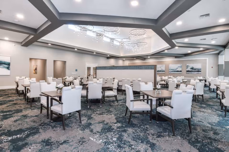Spacious dining room with multiple tables and white upholstered chairs beneath a high coffered ceiling and modern chandeliers.
