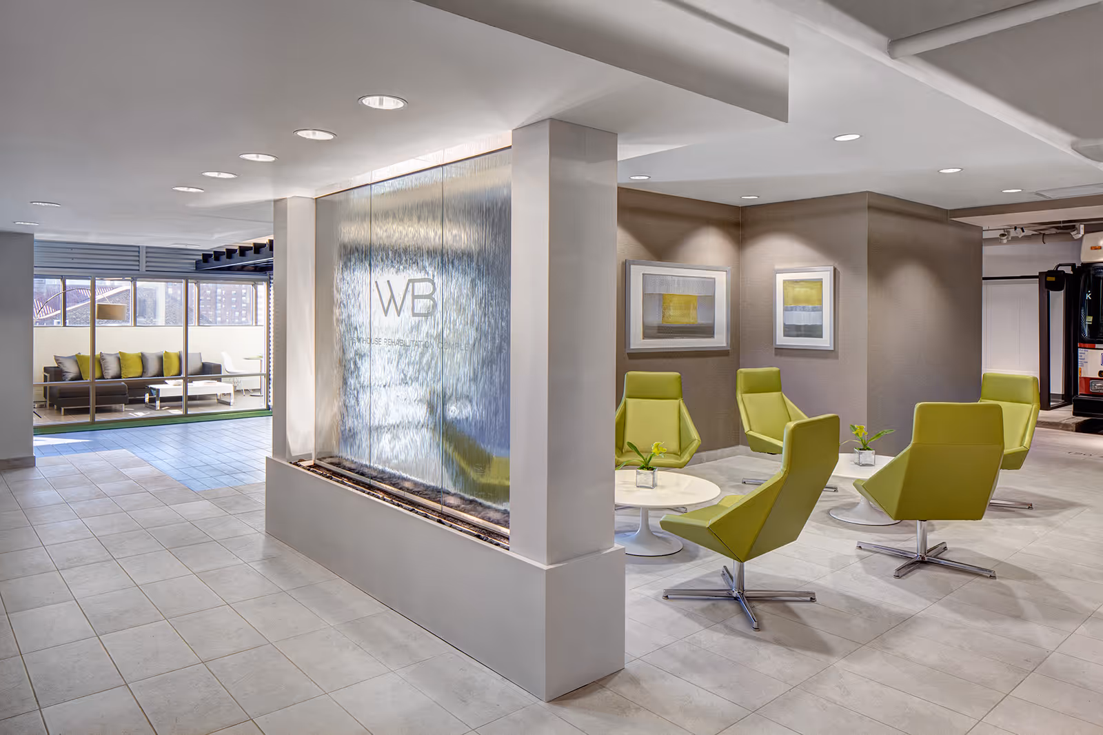 Modern indoor seating area with lime green swivel chairs arranged around white circular tables. A large water feature with the initials 'WB' and the words 'Warren Barr Gold Coast' is prominently displayed. The space is well-lit with recessed ceiling lights and has a tiled floor. In the background, there is an outdoor seating area with a sofa and cushions visible through large glass windows.