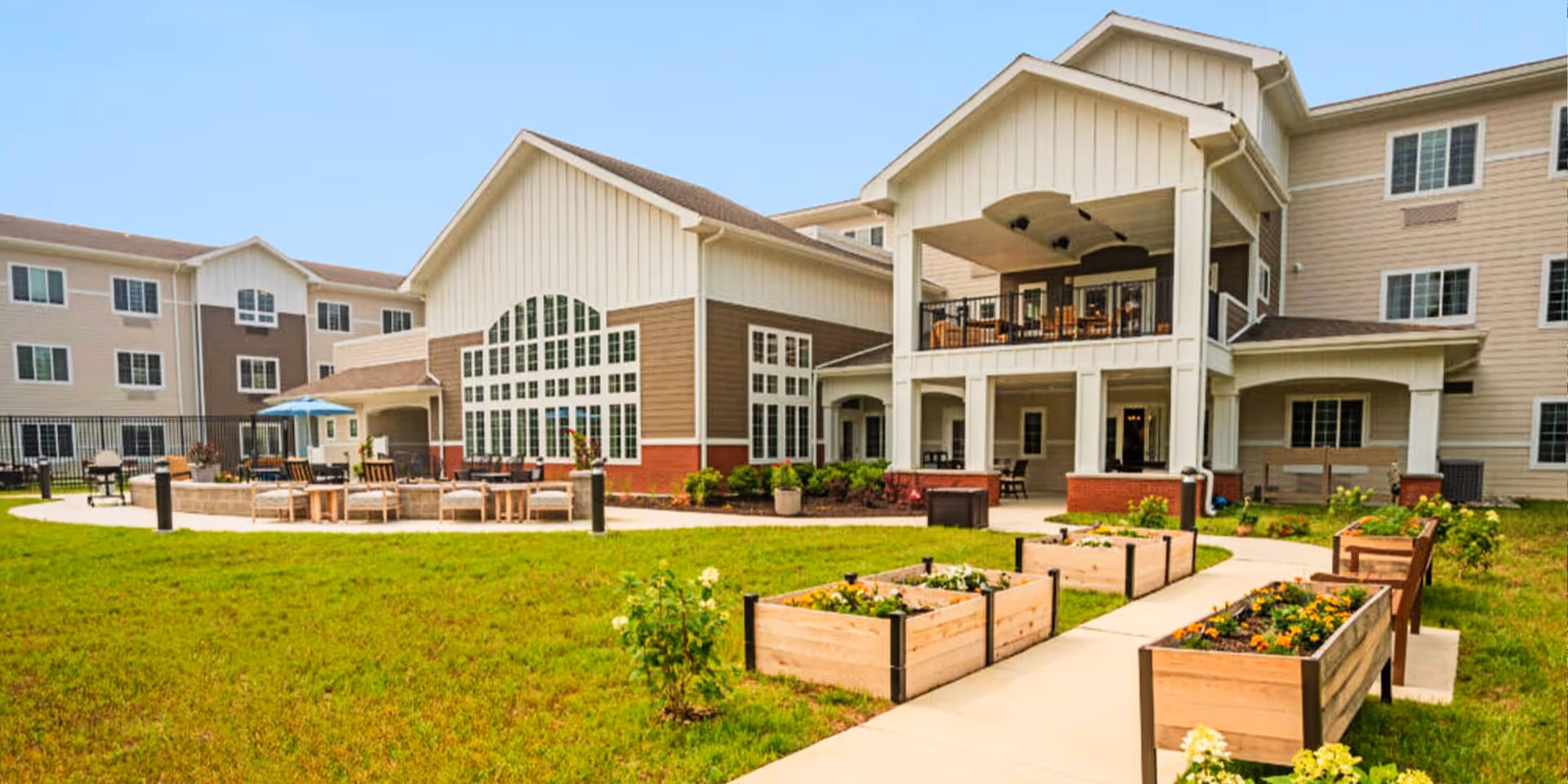 Exterior view of Juniper Village at Newark showing a multi-story senior living building with large windows, a covered balcony with seating, a patio area with chairs and tables, and raised garden beds with flowers along a paved walkway.