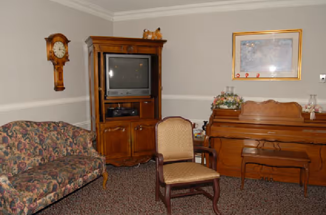 Cozy sitting room with a floral sofa, wooden TV cabinet, upright piano, and upholstered chairs against beige walls.