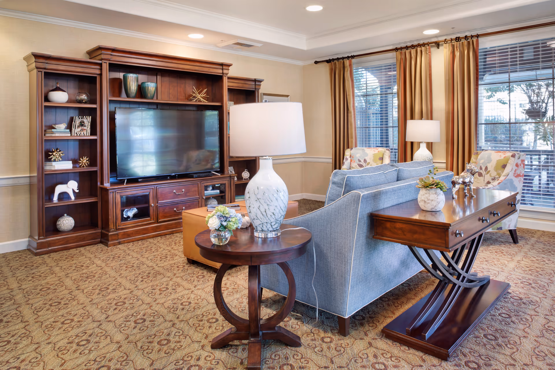 A cozy living room with a blue sofa, wooden entertainment center holding a large TV, side tables with lamps, and large windows with curtains.