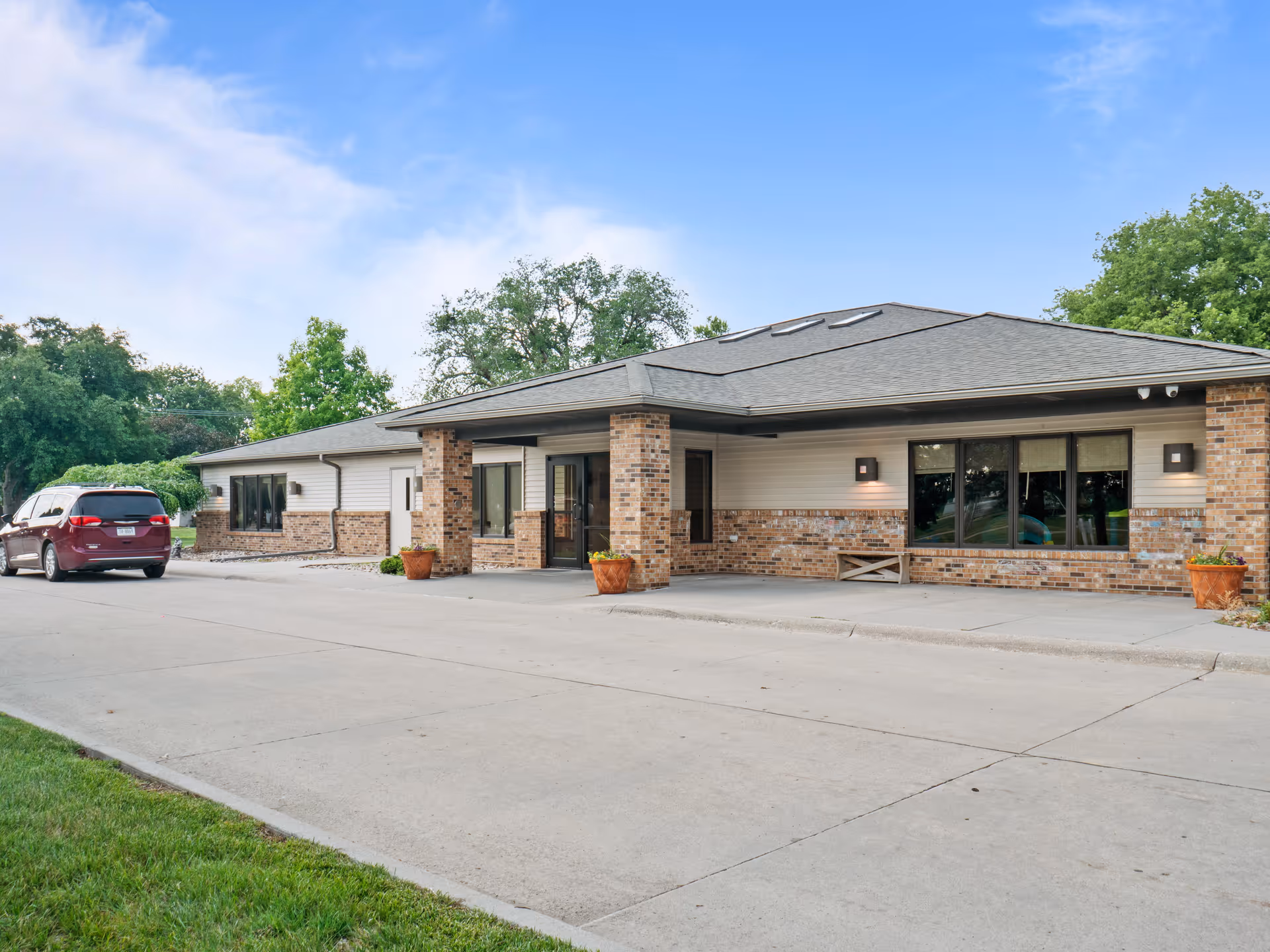 Exterior view of a single-story brick and siding building with a covered entrance, large windows, and a parked maroon minivan on the driveway. The sky is clear with some clouds and there are trees in the background.
