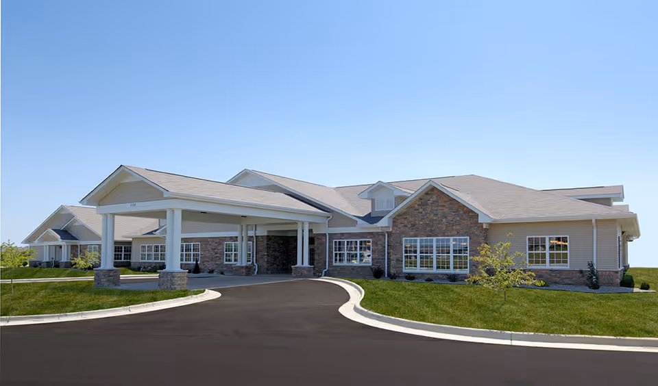 Exterior view of a single-story senior living facility building with a covered entrance, stone and siding facade, multiple windows, and a curved driveway surrounded by green grass under a clear blue sky.