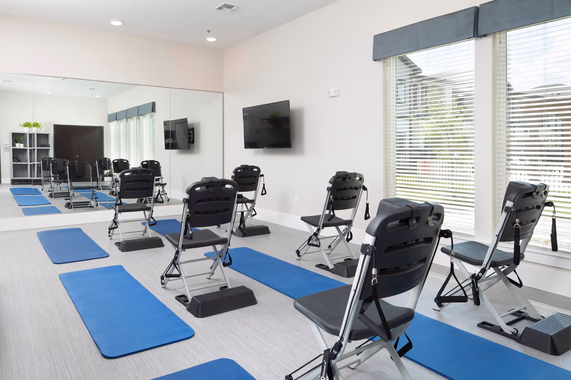A bright exercise room with several black folding chairs equipped with resistance bands, each paired with a blue exercise mat on the floor. The room has large windows with blinds, a wall-mounted TV, a large mirror covering one wall, and a shelving unit with plants and towels.