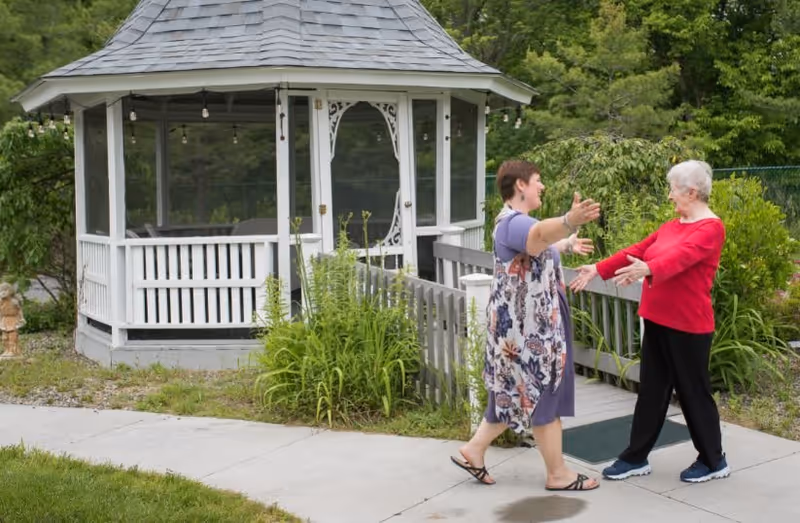 Two women greeting each other with open arms on a paved walkway in front of a white gazebo surrounded by greenery.