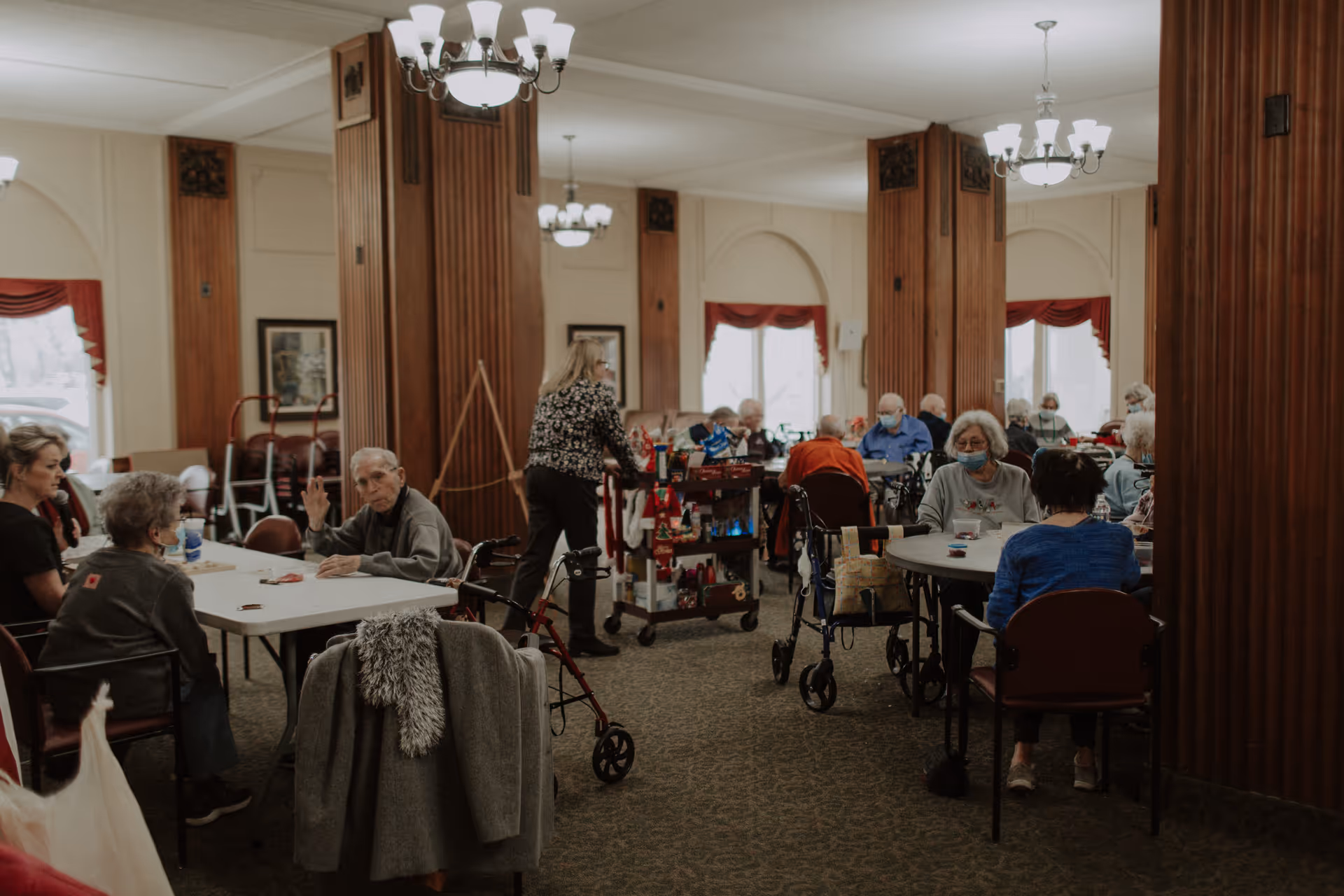 Elderly residents seated at round and rectangular tables in a well-lit communal dining/activity room with staff and walkers.