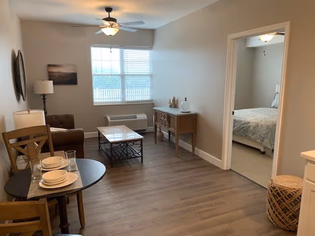 A cozy living room area with a small round dining table set for two, a brown armchair, a coffee table, a sideboard, and a ceiling fan with light. A window with blinds lets in natural light. An open doorway leads to a bedroom with a bed covered in a light-colored bedspread.
