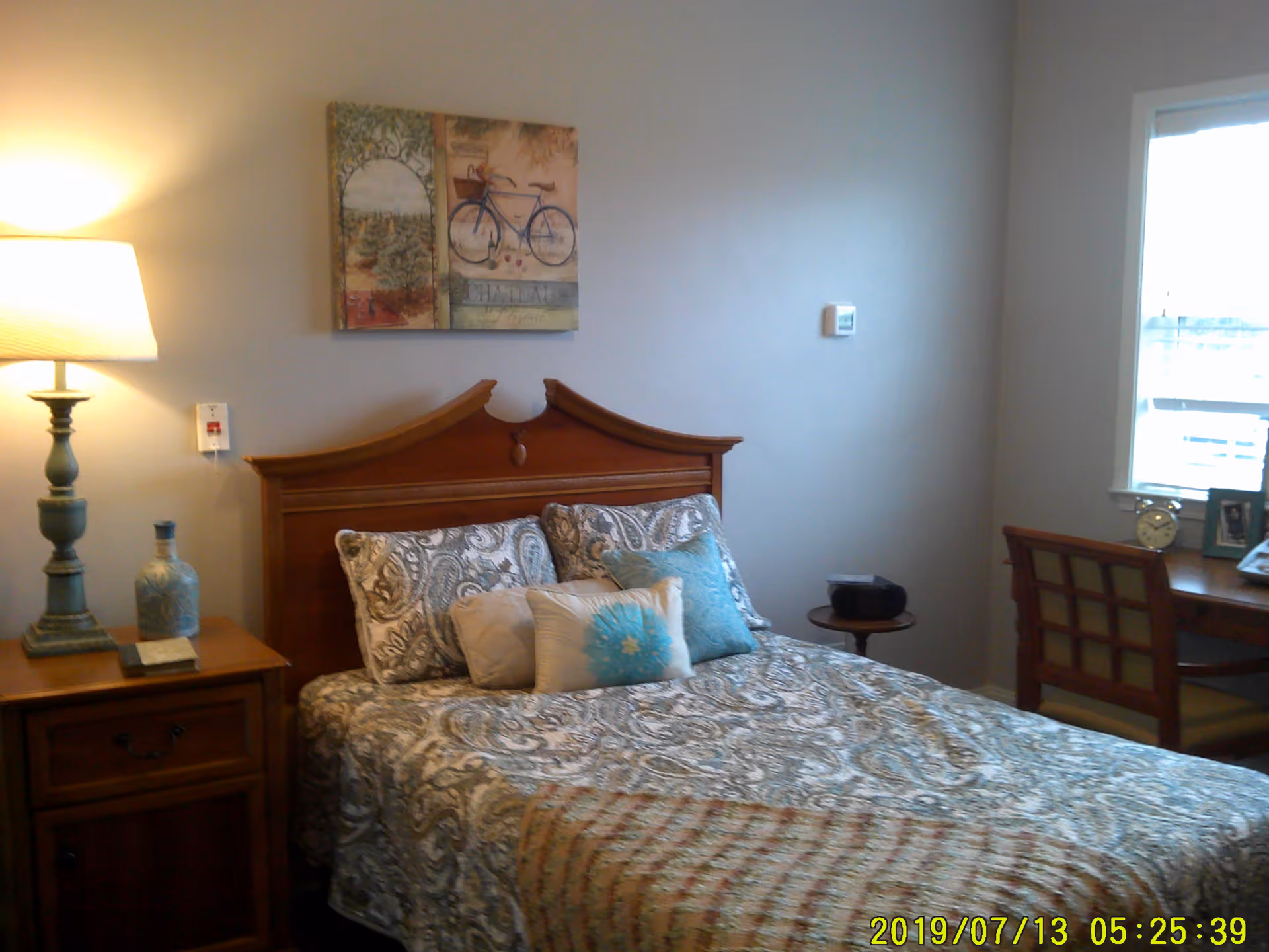 A bedroom with a wooden headboard bed covered in patterned bedding and multiple pillows. To the left of the bed is a wooden nightstand with a lamp and decorative items. On the wall above the bed is a painting featuring a bicycle. To the right, there is a window with a wooden desk and chair beneath it, holding a clock and framed photo.