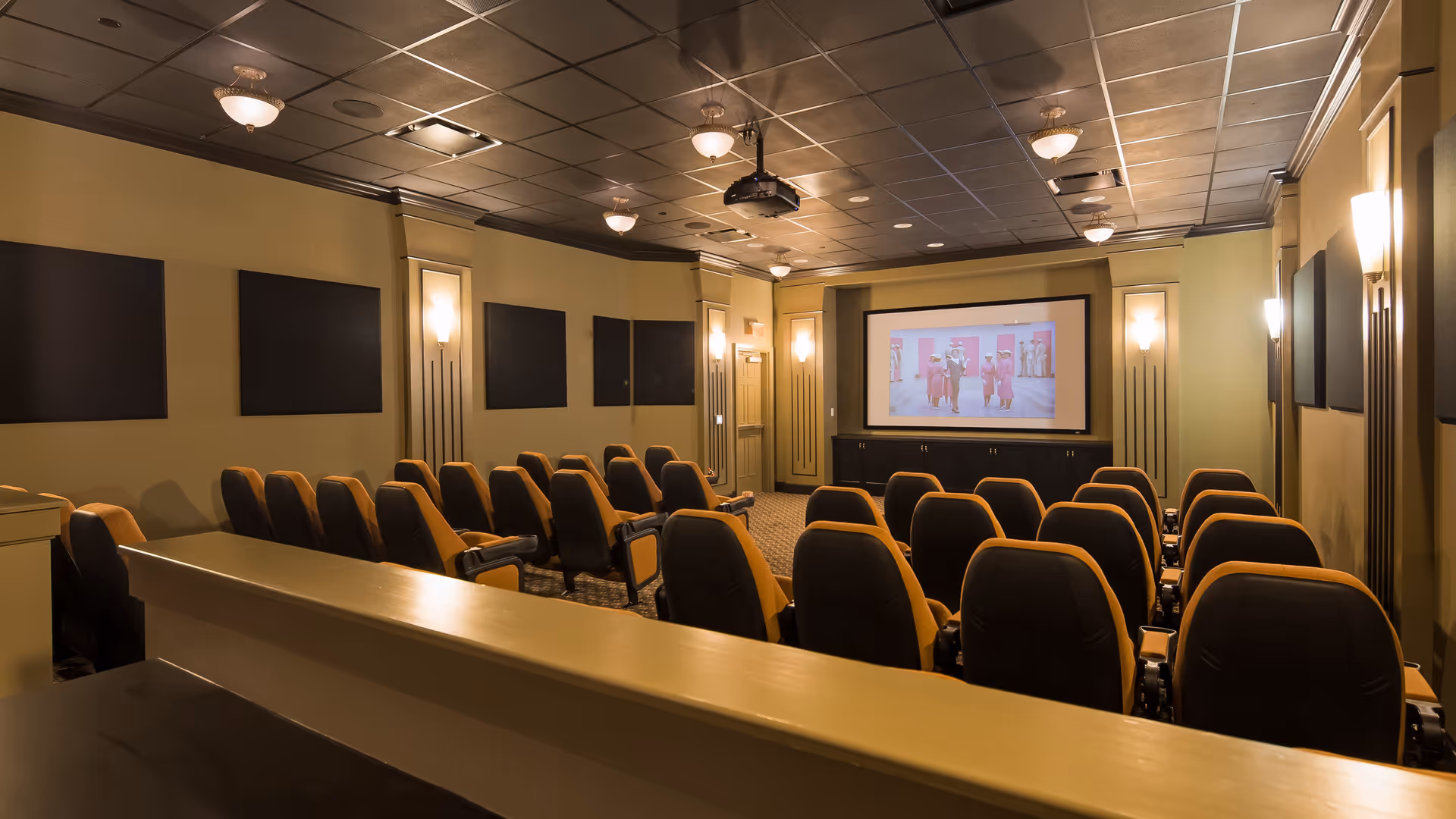 Interior view of a small theater room with rows of black and tan cushioned seats facing a large screen displaying a movie scene. The room has beige walls with black acoustic panels and soft wall lighting, and a ceiling-mounted projector.