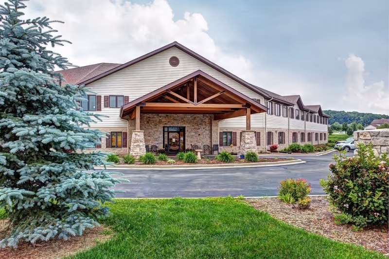 Front exterior of a two-story senior living building with a covered wooden entrance, stone facade, driveway, and landscaped lawn.