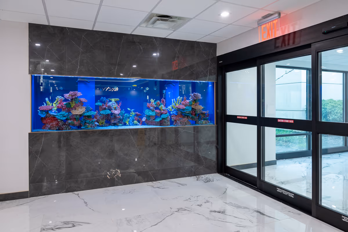 Interior view of a hallway featuring a large built-in aquarium with colorful coral and fish, next to a glass automatic sliding door with an illuminated red EXIT sign above it. The floor is white marble with gray veining, and the walls around the aquarium are covered in dark marble tiles.