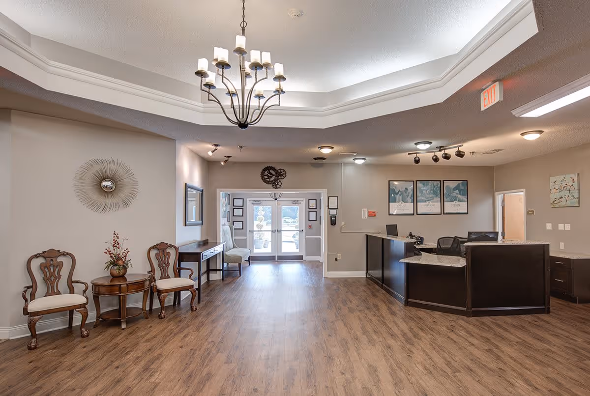 A spacious senior living facility lobby with wooden flooring, a chandelier hanging from a tray ceiling, two wooden chairs with cushions and a small round table with a flower vase on the left, a reception desk on the right, and glass double doors at the far end letting in natural light.