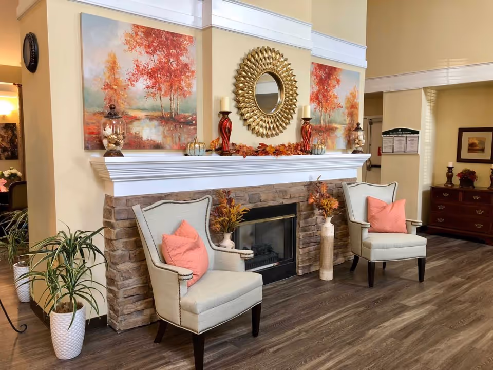 Cozy senior living common area with a stone fireplace mantel decorated for autumn and two upholstered armchairs with coral pillows.