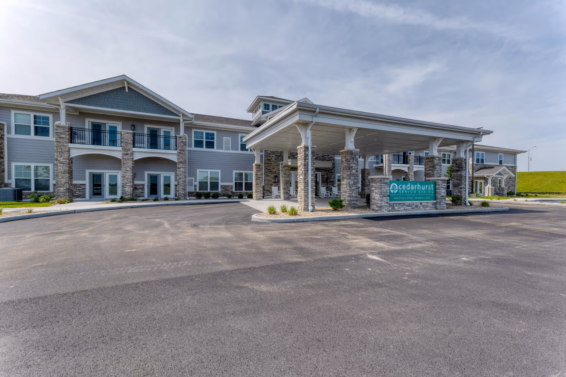 Exterior view of Cedarhurst Senior Living of Dyer building with a covered entrance supported by stone pillars, multiple windows, and a large paved driveway under a partly cloudy sky.