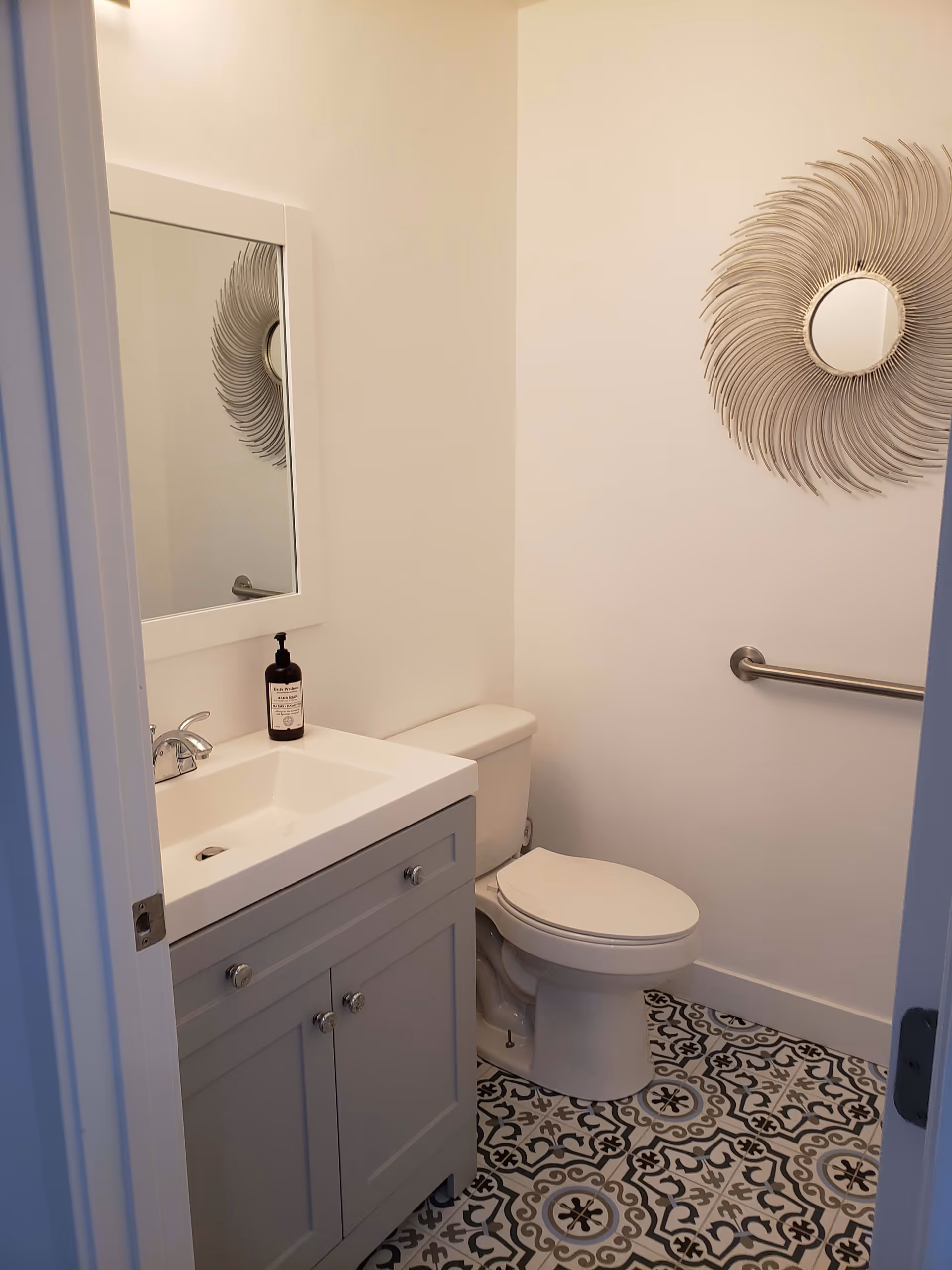 A small bathroom with a white toilet and a gray vanity with a white sink. Above the sink is a rectangular mirror with a white frame. On the wall beside the toilet is a decorative round mirror with a sunburst design and a metal grab bar. The floor has patterned tiles in black, white, and gray.