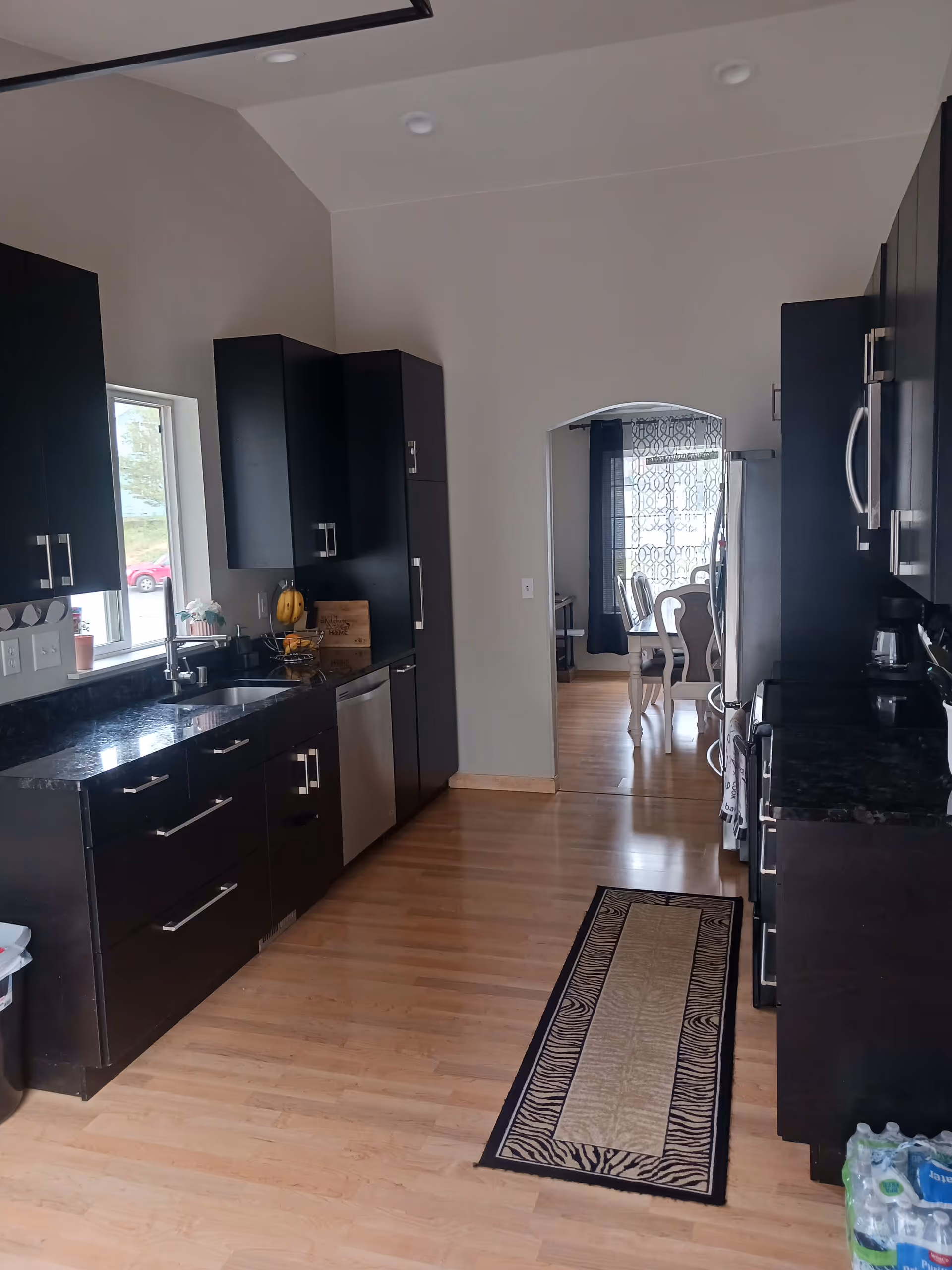 A modern kitchen with dark cabinetry and stainless steel appliances including a dishwasher and refrigerator. The kitchen has a light wooden floor and a patterned runner rug. There is a window above the sink letting in natural light. Through an arched doorway, a dining area with white chairs and a window with patterned curtains is visible.