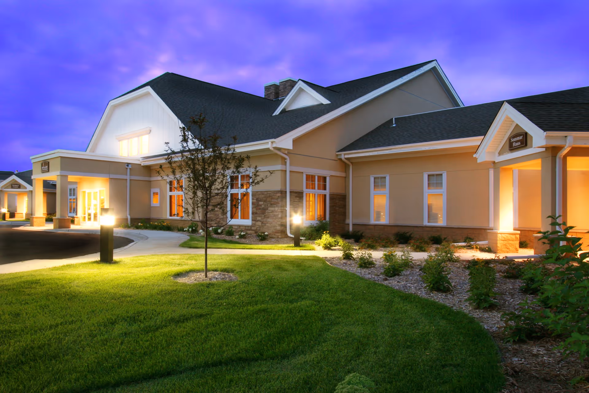 Front exterior of a well-lit senior living facility building with landscaped lawn at dusk.