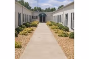 Concrete walkway flanked by low shrubs and two single-story building wings leading to an arched entrance with a green awning.