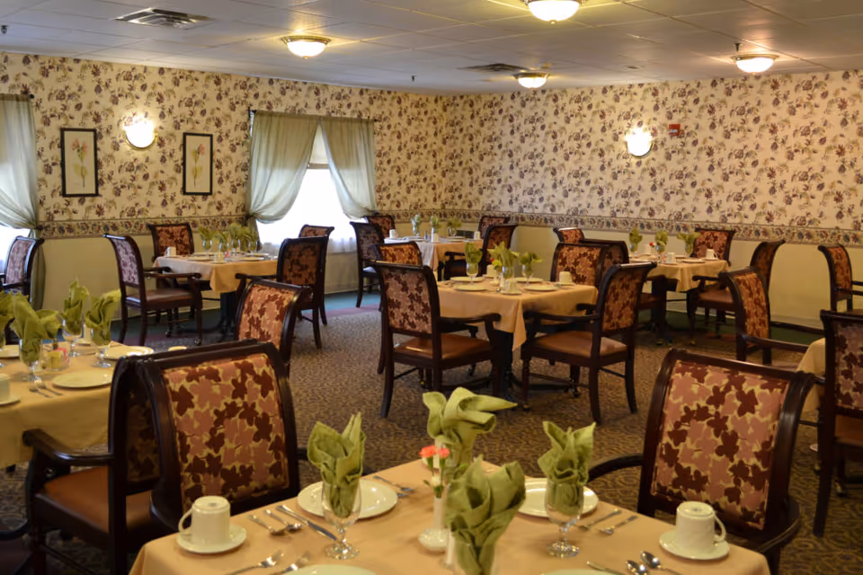 Dining room with multiple tables set with plates, cups and folded green napkins, floral-upholstered chairs and floral wallpapered walls.